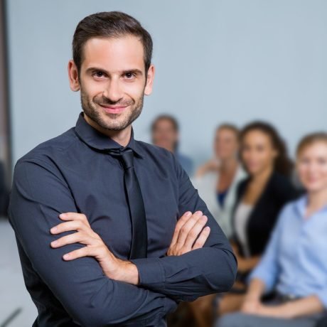 Portrait of successful business leader standing with crossed arms and smiling, business people sitting at office desk on background