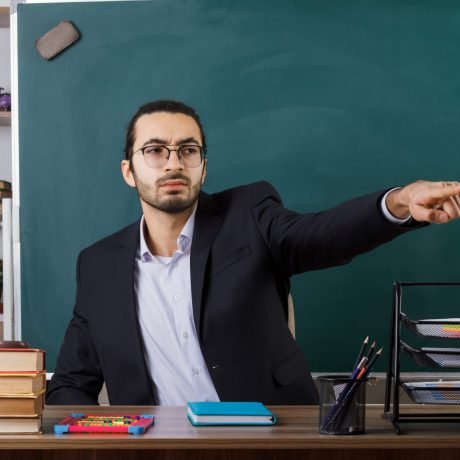 strict points at side male teacher wearing glasses sitting at table with school tools in classroom