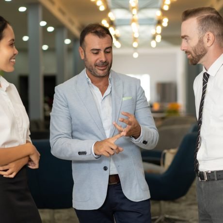 Three positive business people talking in office lobby. Businesspeople standing and discussing idea with blurred interior in background. Business coworkers and communication concept.